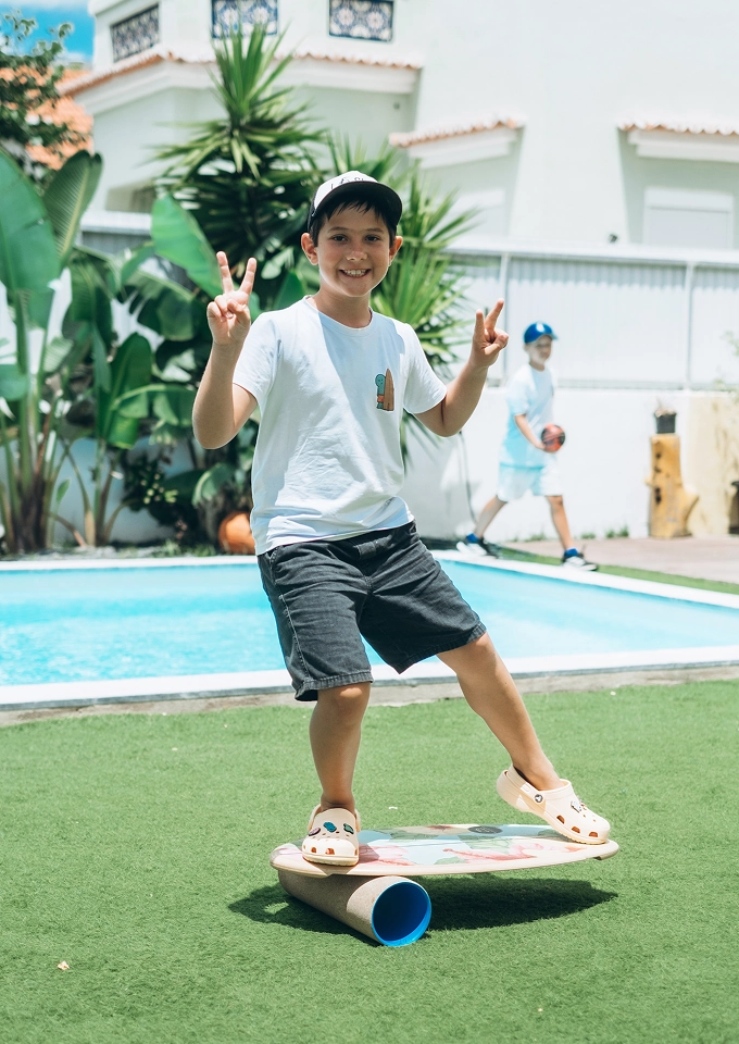Boy balancing on a board over a cylindrical roller on grass near a pool, making peace signs with both hands.