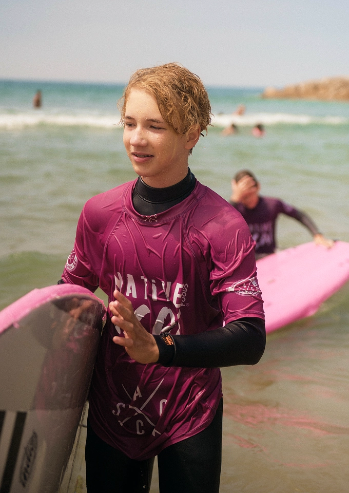 Teen boy with wet blond hair wearing a purple rash guard holding a surfboard on the beach.