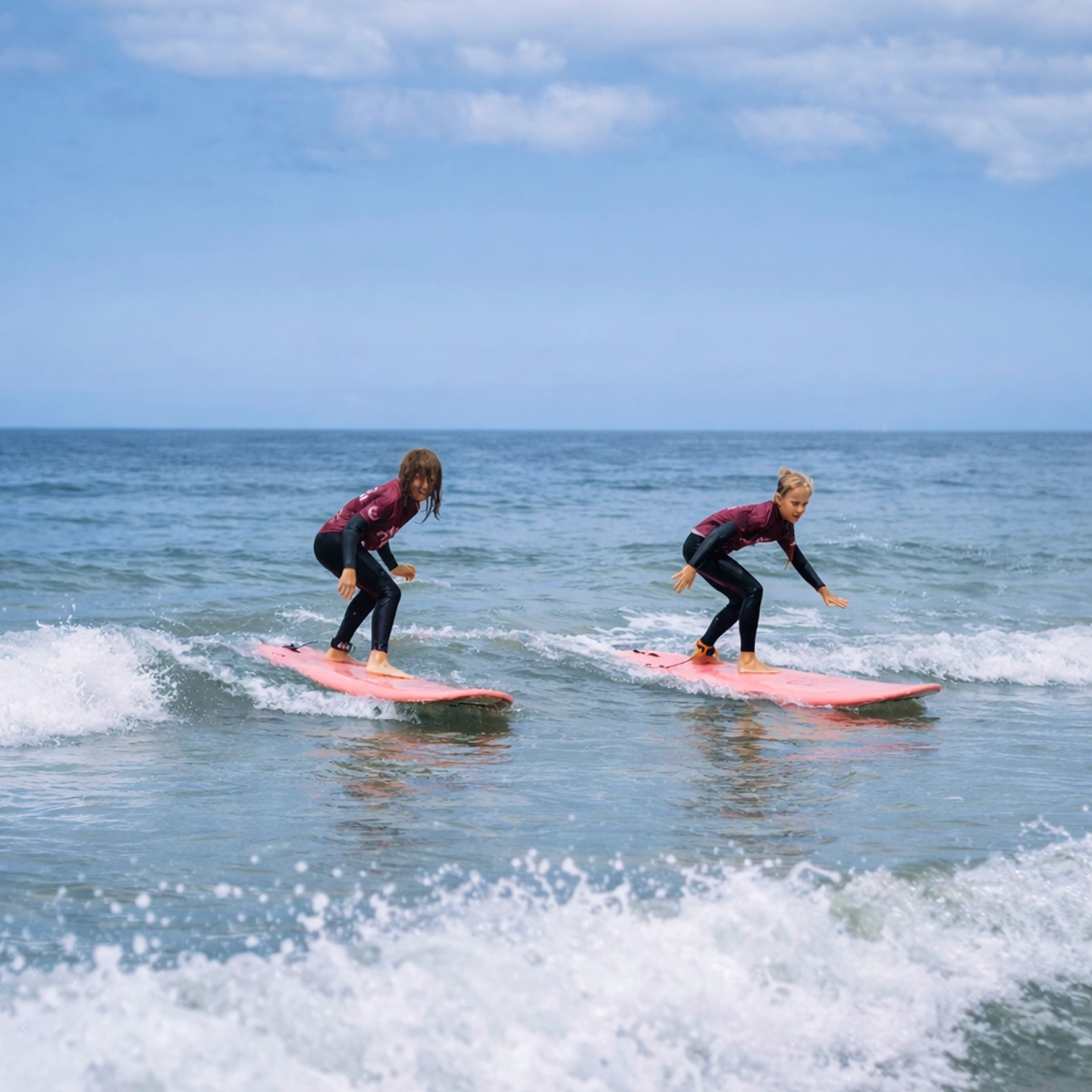 Two children surfing on small waves on pink surfboards wearing wetsuits and maroon rash guards.
