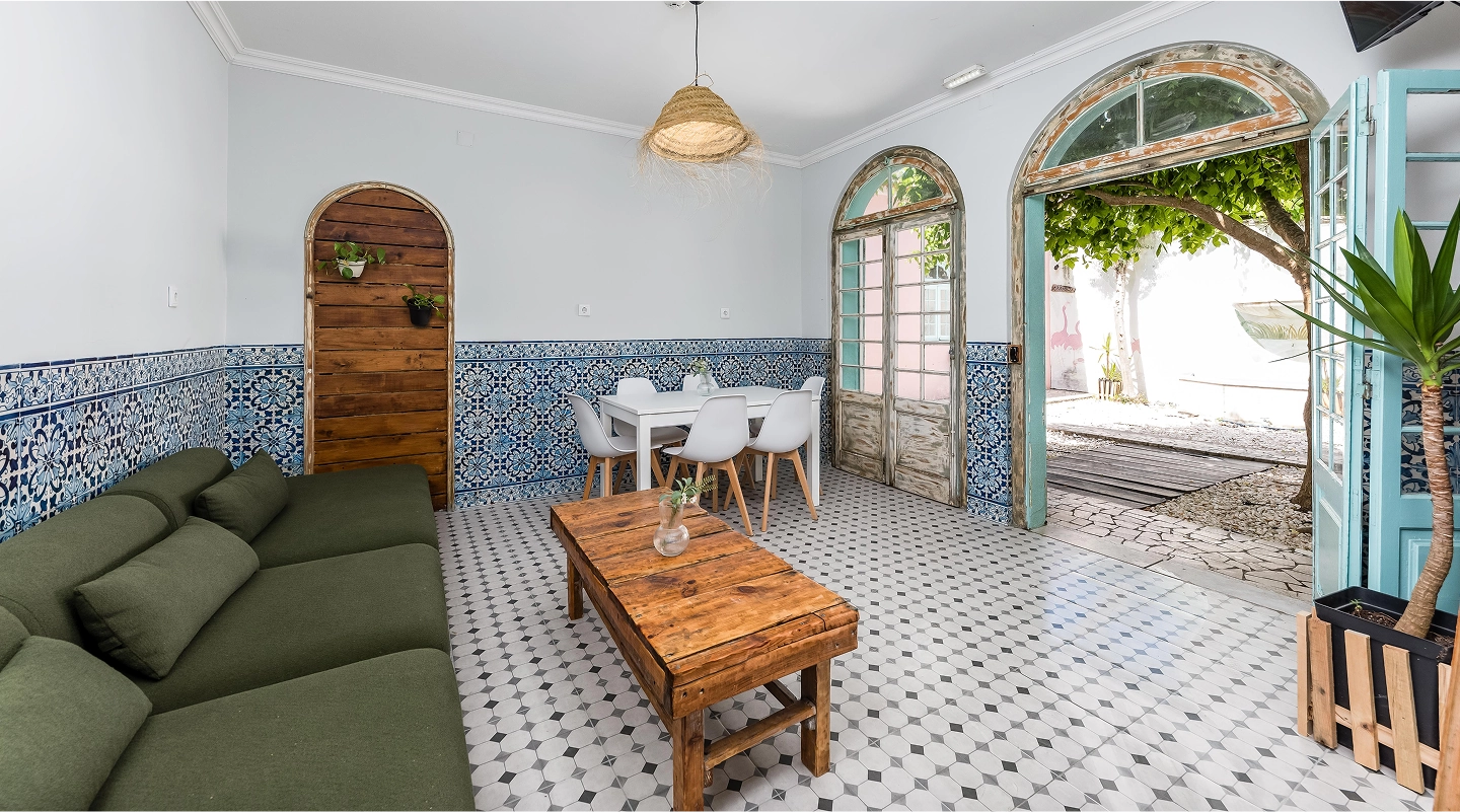 Bright room with green sofa, wooden table, white dining set, blue patterned tiles, and open arched doors to garden.