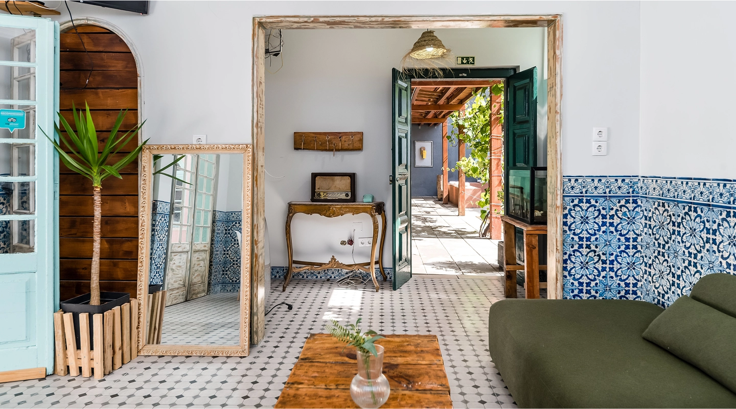 Bright room with patterned floor tiles, large mirror, green sofa, wooden table, and open door leading to a sunlit patio.