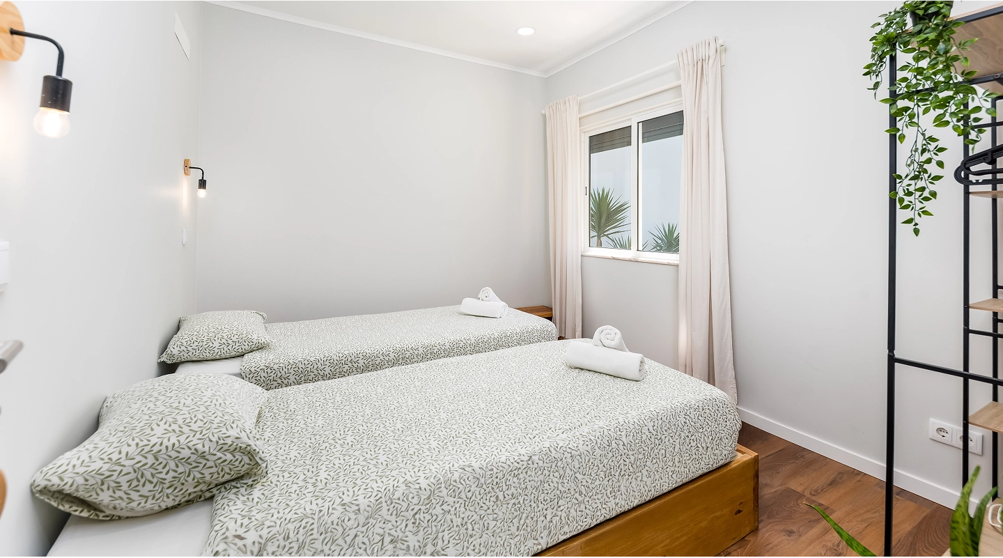Minimalist bedroom with two single beds with patterned bedding and a window with cream curtains.