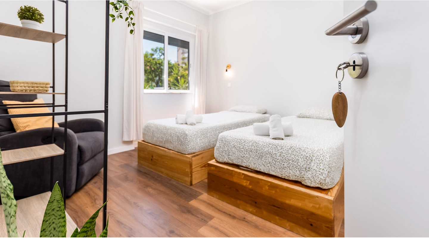 Modern bedroom with two single beds, white bedding, wooden bed frames, and a window showing greenery.