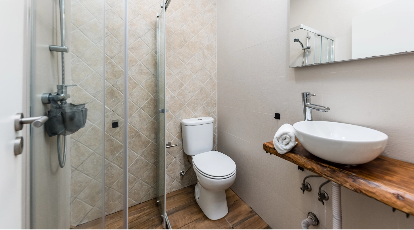 Small bathroom with white toilet, glass shower enclosure, wooden sink countertop, and round white basin with towel.