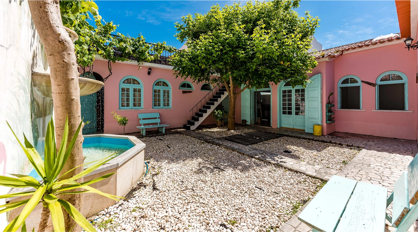 Courtyard with pink walls, turquoise windows and doors, white stone ground, a fountain, and a leafy tree.