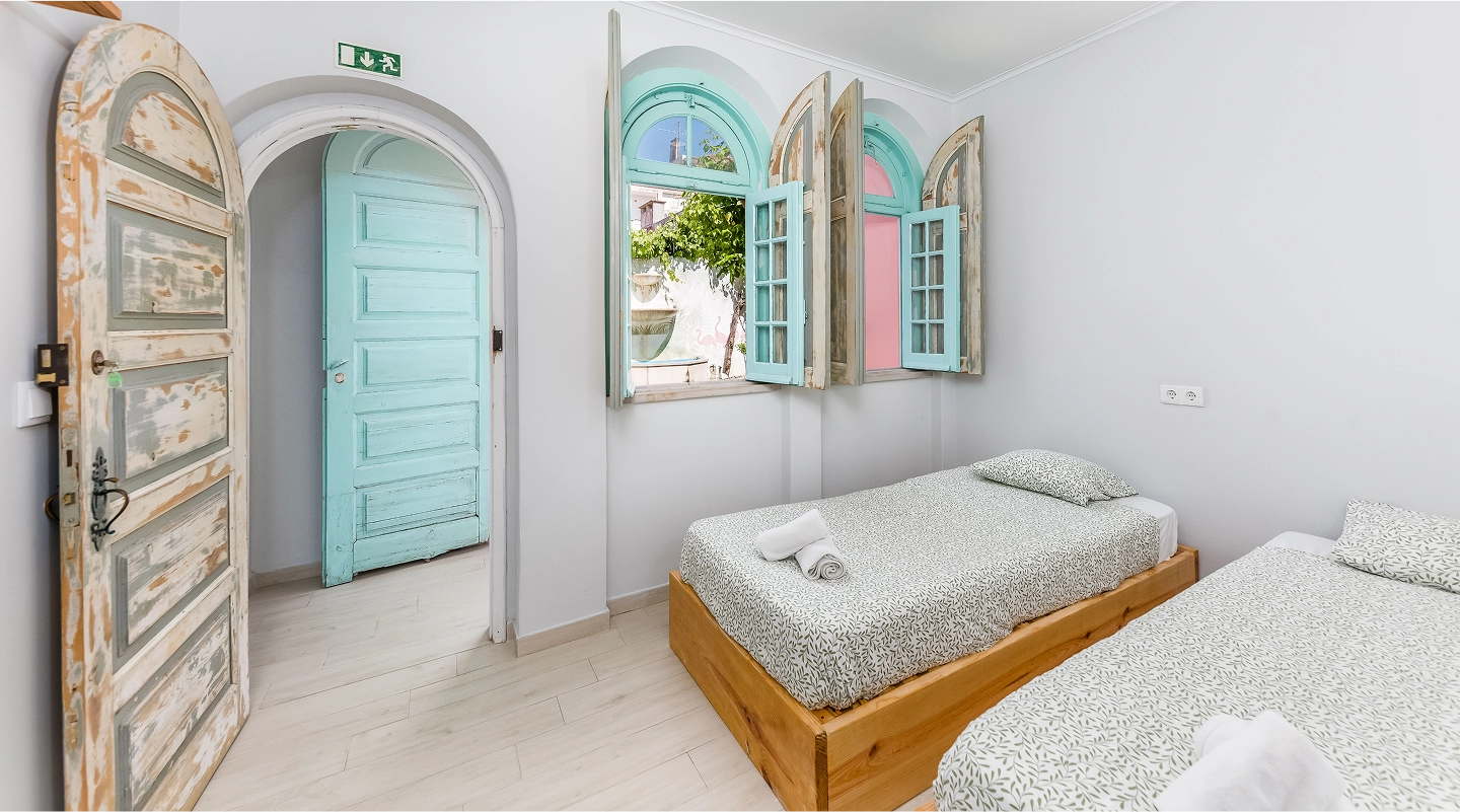 Minimalist bedroom with two single wooden beds, patterned bedding, and vintage turquoise window shutters open to a garden.