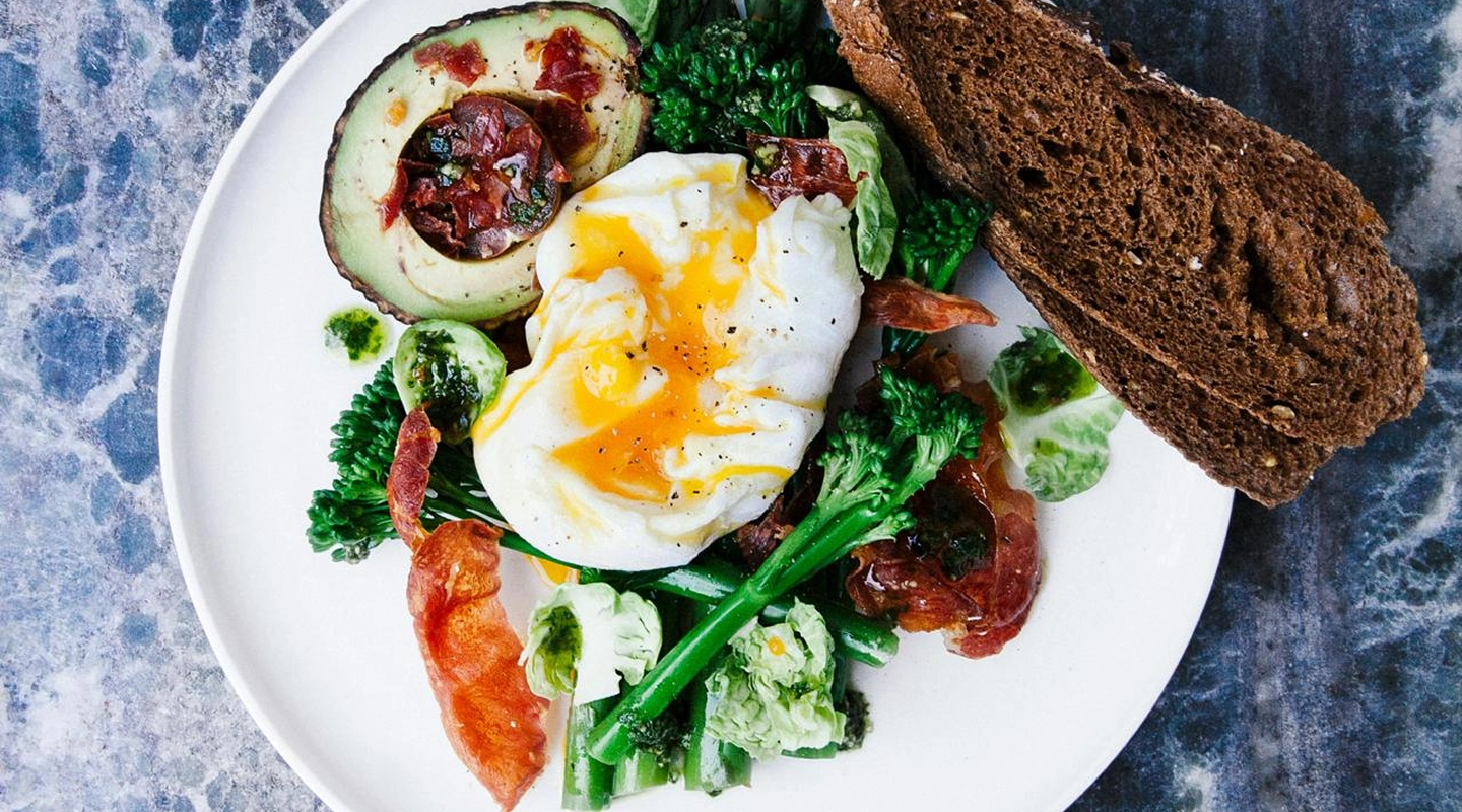 Poached egg with avocado, broccolini, greens, crispy bacon, and two slices of brown bread on a white plate.