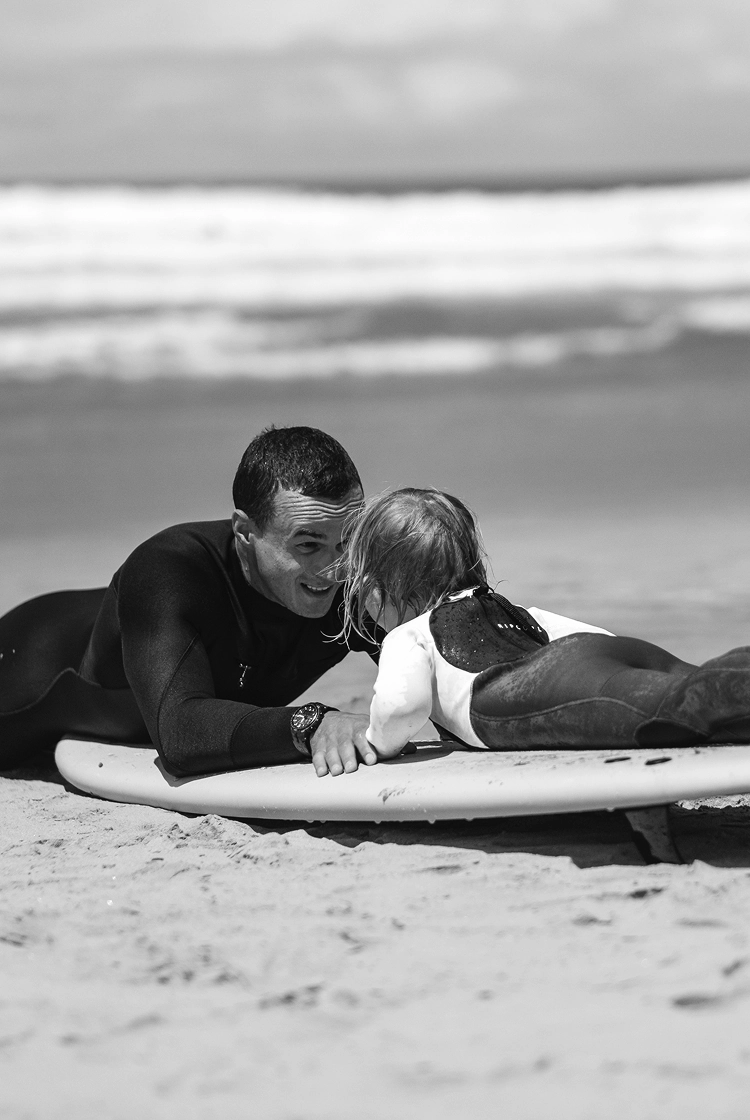 Man and child in wetsuits lying on a surfboard on the beach, facing each other and smiling.